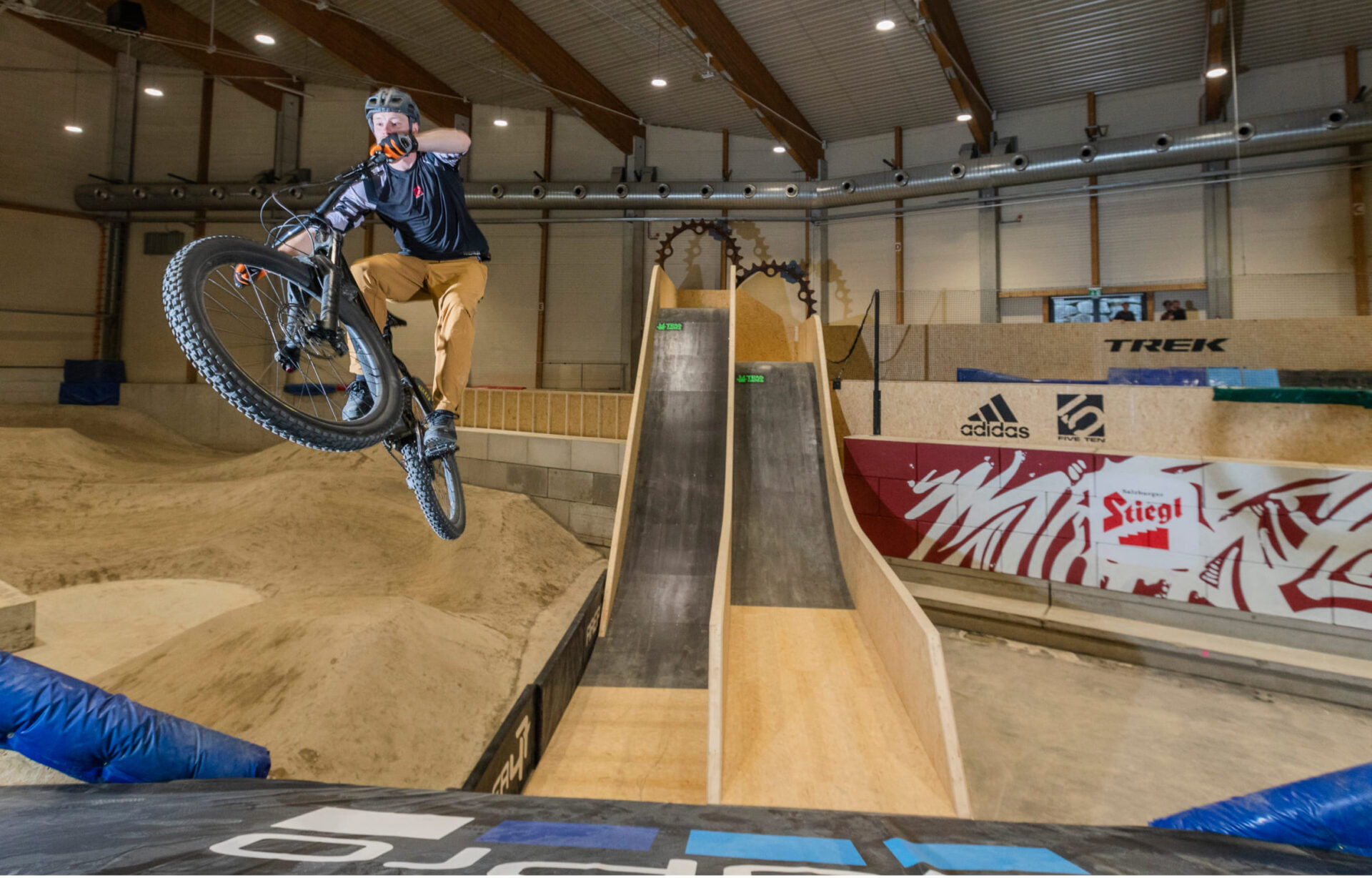 Mountain bikers jump from a wooden ramp in the AREA 47 Indoor Bike Park in Ötztal, Tyrol
