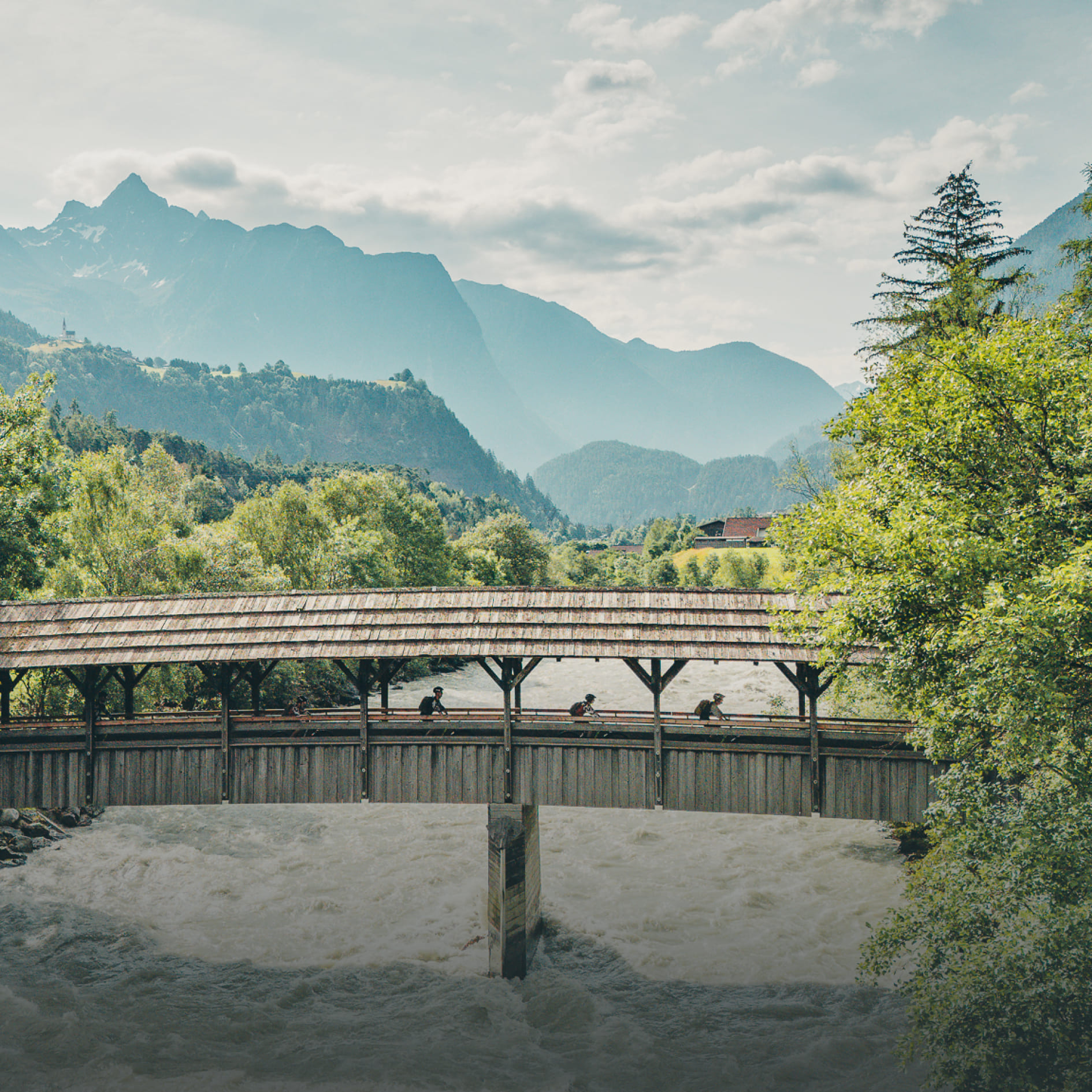 Radfahrer überqueren eine überdachte Holzbrücke in der AREA 47 im Ötztal, Tirol, mit Bergpanorama im Hintergrund