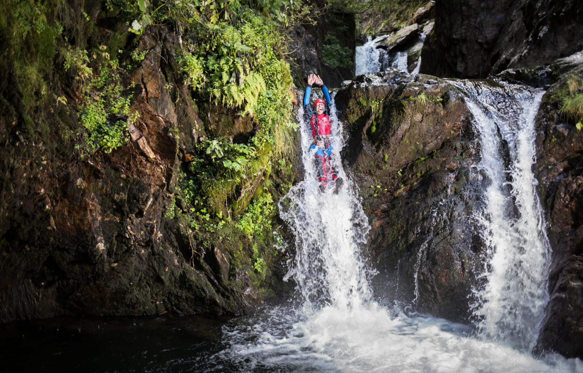 Person in roter Canyoning-Ausrüstung rutscht durch einen Wasserfall in einer Schlucht in der AREA 47 im Ötztal, Tirol.