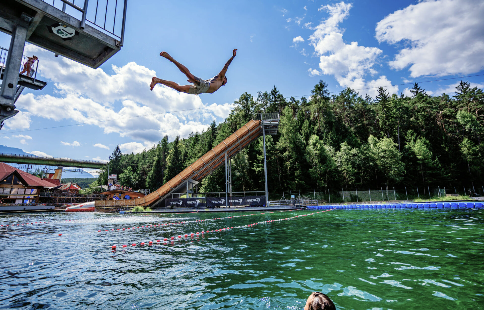Mann springt von einem hohen Turm ins Wasser in der Water AREA der AREA 47, umgeben von Wald und blauen Himmel