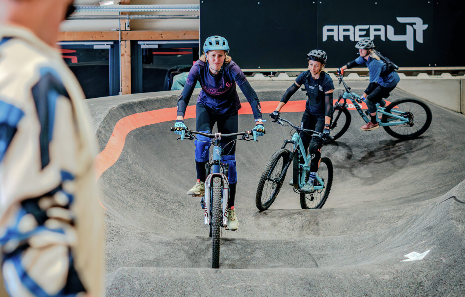 Frau fährt durch den Pumptrack im Indoor-Bikepark der AREA 47, gefolgt von weiteren Teilnehmern