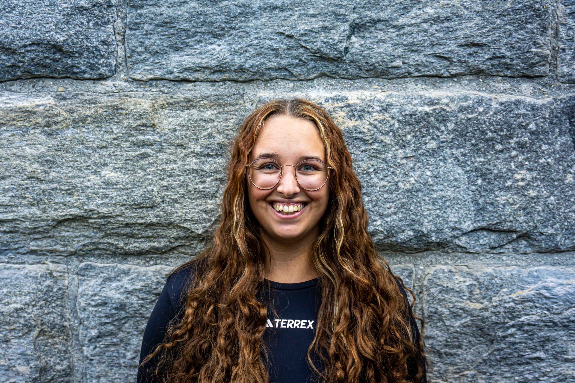 Portrait of a female staff member with long curly hair and glasses standing in front of a stone wall at AREA 47 in the Ötztal, Tyrol