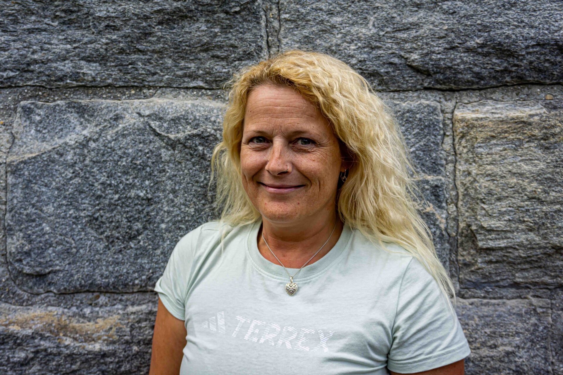 Porträt einer Mitarbeiterin mit blondem Haar im hellen TERREX-Shirt vor einer Steinwand in der AREA 47 im Ötztal, Tirol