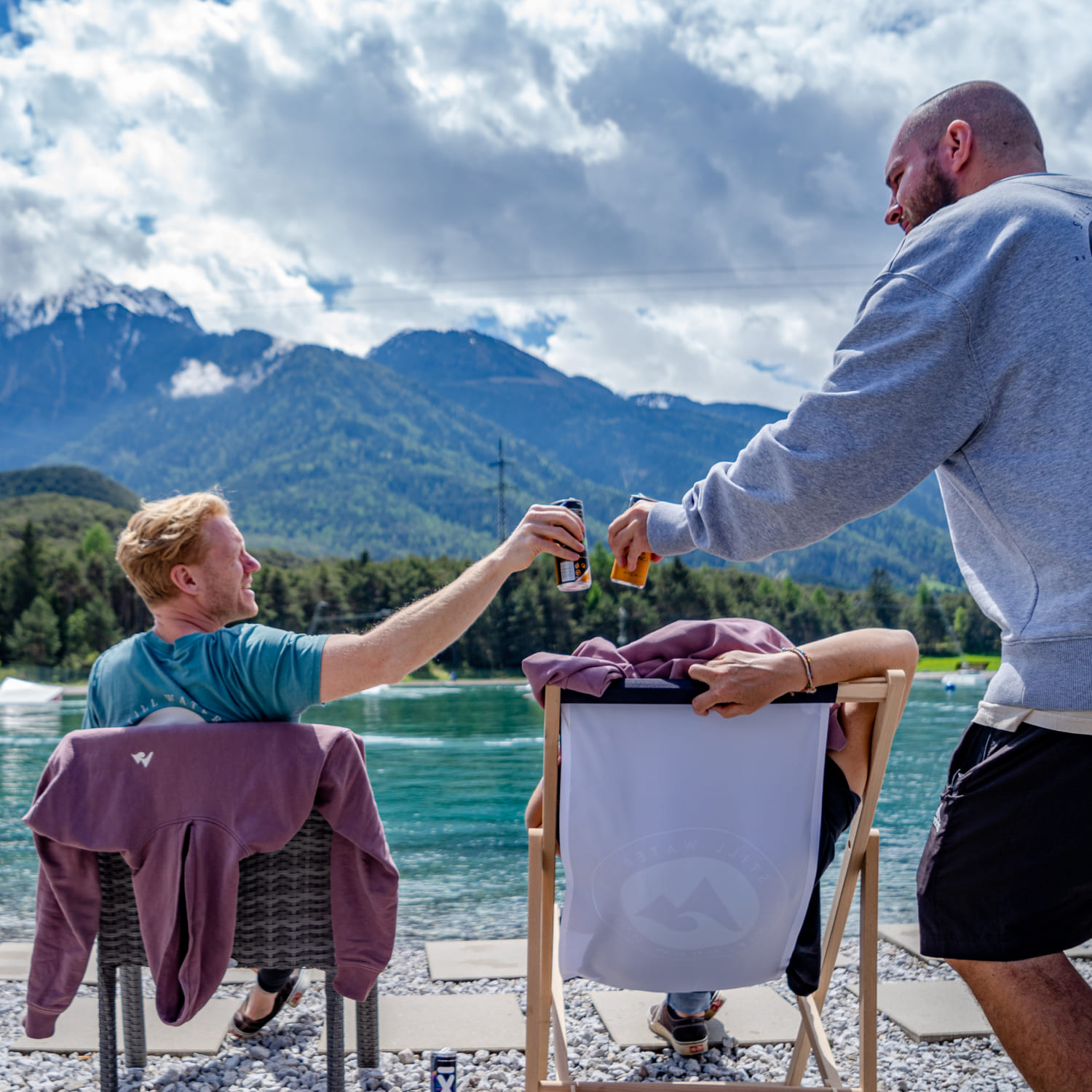 Friends in the Wake Café at AREA 47 toast with drinks in front of the Water AREA in Ötztal, Tyrol